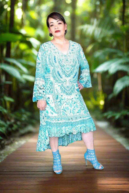 Woman wearing peacock silk hi-low frill dress with long sleeves and intricate beadwork standing on wooden pathway in lush garden