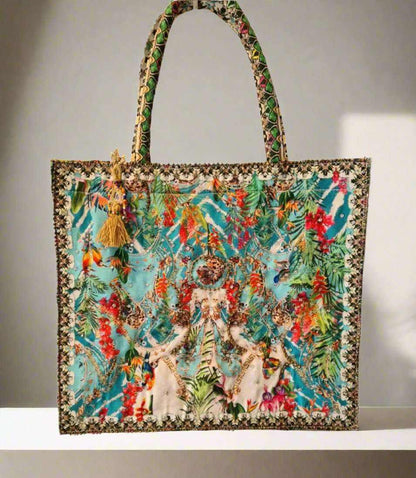 Large embellished tote bag with colorful floral and nature-inspired print, padded handles, zipper closure, and tassel accents