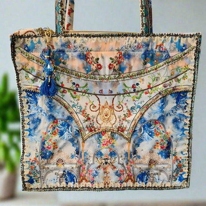 Large embellished tote bag with colorful floral and architectural print, padded handles, and zipper closure